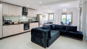 A modern kitchen with white cabinets, a black countertop, and stainless steel appliances. There is a dark blue sofa and a dining area with a view of the sea. - The perfect coastal getaway, just steps from the beach, with a private balcony, open living area. (Tangier)