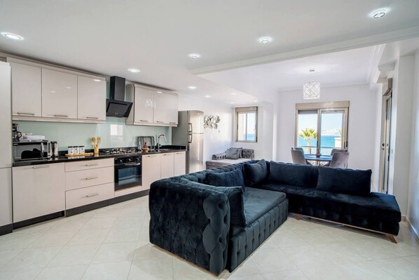 A modern kitchen with white cabinets, a black countertop, and stainless steel appliances. There is a dark blue sofa and a dining area with a view of the sea.