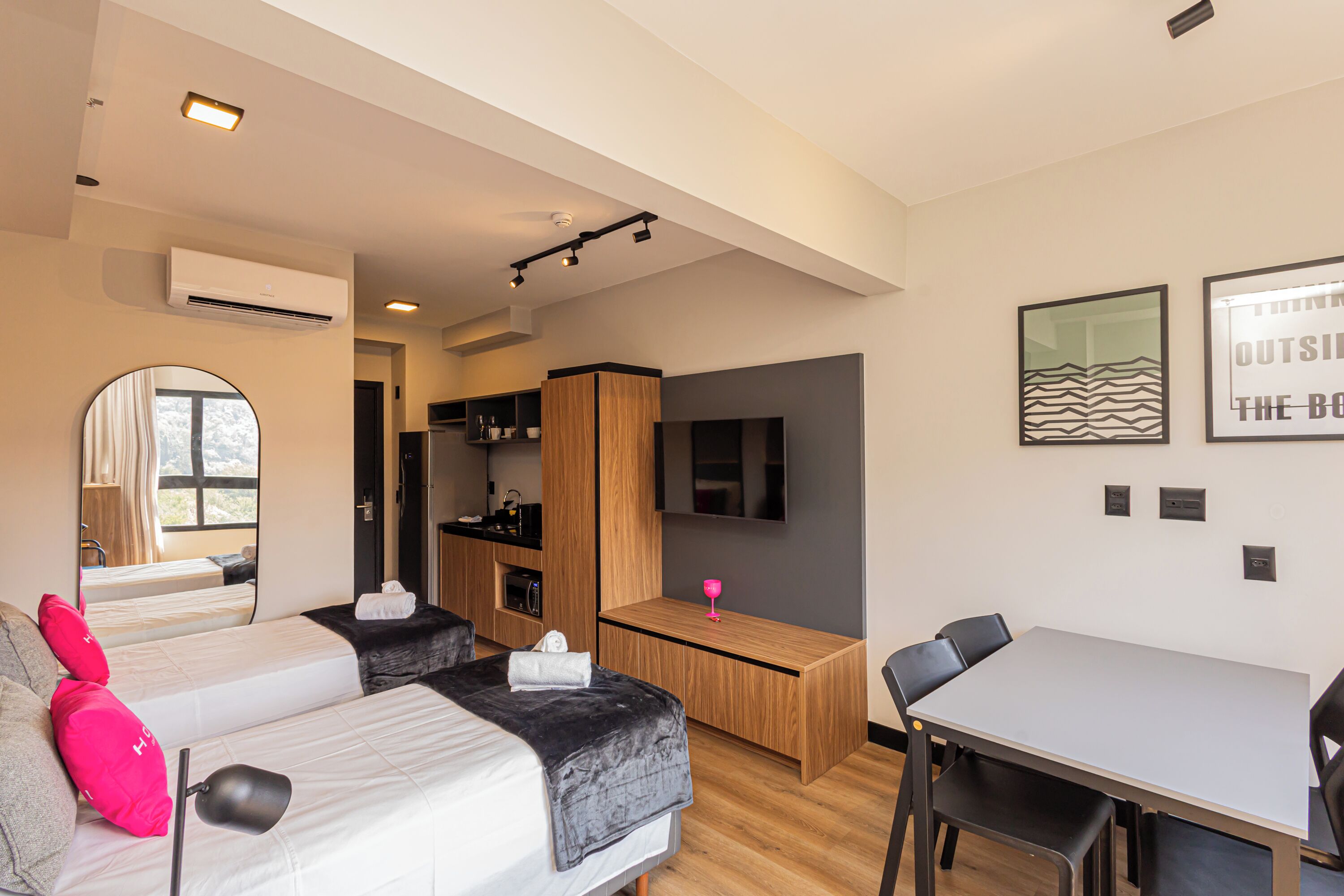 A modern hotel room with two beds, a dining table, a TV, and a kitchen area.