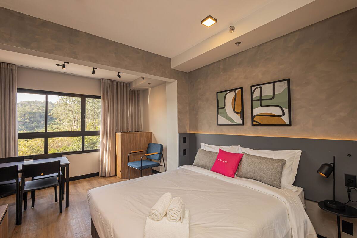 Une chambre d’hôtel moderne avec un grand lit, un bureau et une chaise. On y trouve une fenêtre avec des rideaux, des œuvres d’art au mur et une table de chevet avec une lampe.