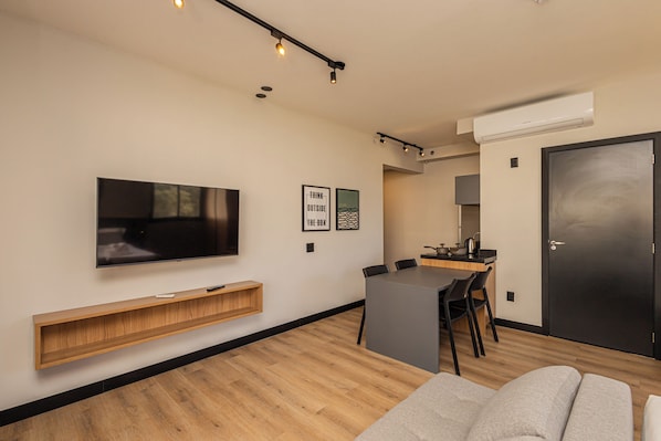 Una sala de estar moderna con un televisor de pantalla plana, una mesa consola de madera, un sofá gris y un comedor con una cocina al fondo.