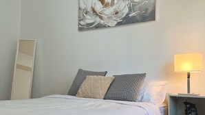 Una cama bien tendida con ropa blanca, un cojín de pata de gallo y un cojín con estampado floral. Se ve una mesita de noche con una lámpara y un pequeño estante.