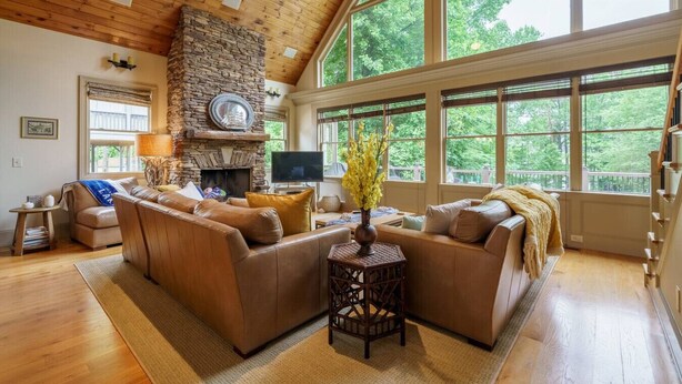 A spacious living room with a stone fireplace, leather sofas, a television, and large windows offering a view of the outdoors. - Beautiful Lake Home/Private Dock (Gainesville)