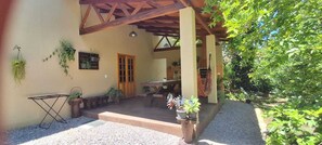 A covered patio area with a wooden table and chairs, potted plants, and a gravel pathway leading to a wooden door. - Beautiful House! Enjoy nature and cool off in the pool. (Morretes)