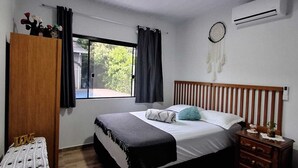 A bedroom with a bed, a wardrobe, a bedside table, a nightstand, a window with curtains, and a wall-mounted air conditioner.