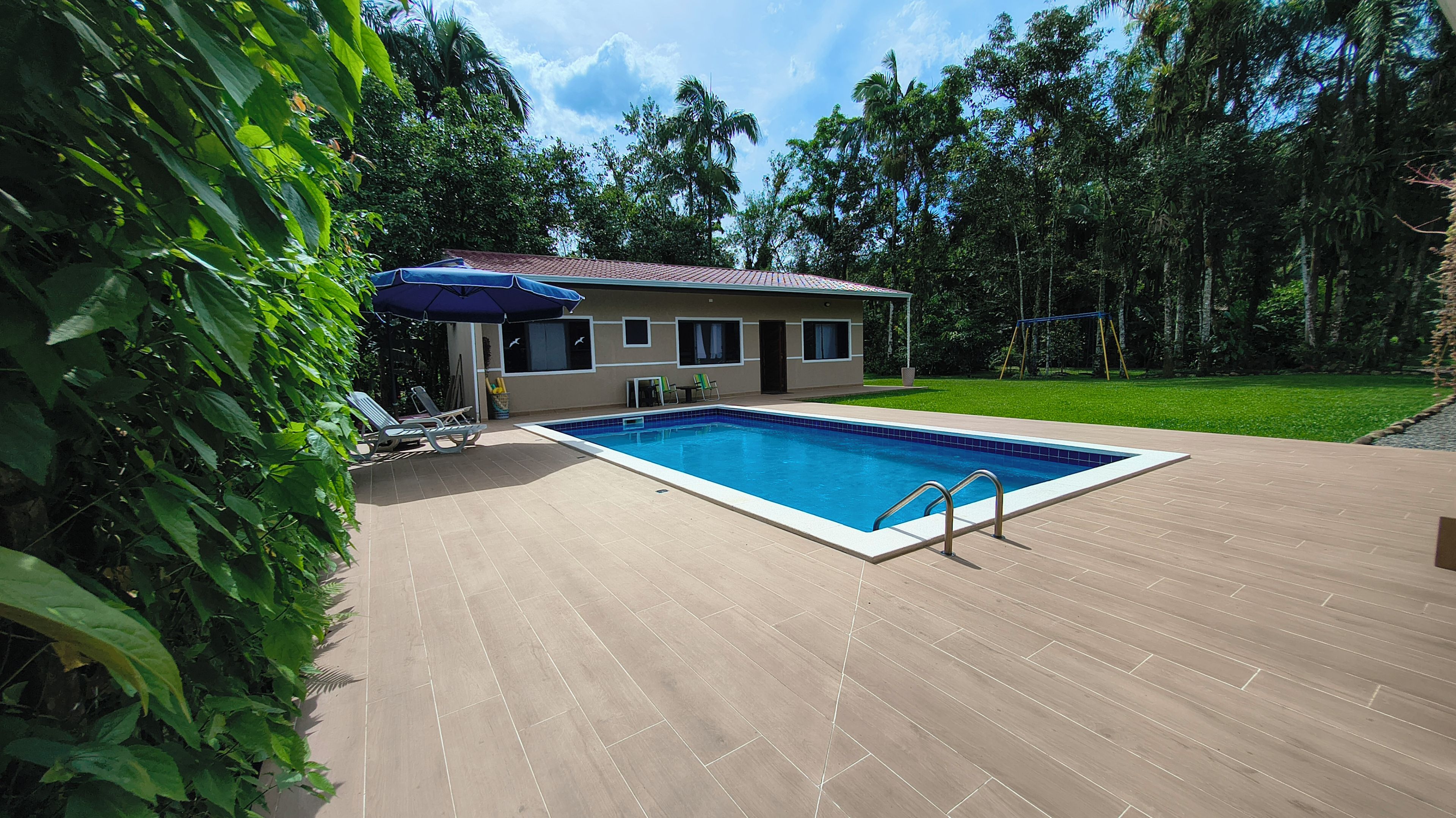 Ein Schwimmbecken mit Fliesenveranda, ein überdachter Sitzbereich mit Stühlen und ein Haus im Hintergrund, umgeben von Bäumen.