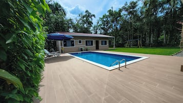 Ein Schwimmbecken mit Fliesenveranda, ein überdachter Sitzbereich mit Stühlen und ein Haus im Hintergrund, umgeben von Bäumen.