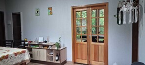 A room with a bed, a wooden desk with a plant, a wine bottle, and a door with a view of greenery.