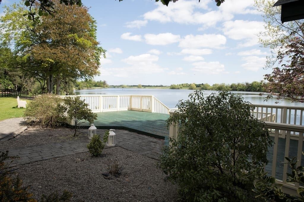 A lakeside deck with a wooden railing, surrounded by trees and shrubs.