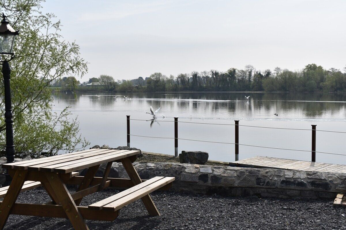 A wooden picnic table by a lake with a stone wall and a lamp post.