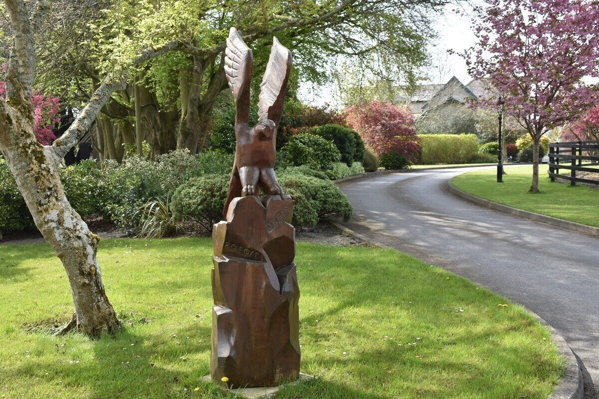 Een beeld van een beer met vleugels in een grasrijk gebied, met een bochtige weg en een huis op de achtergrond.