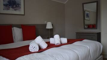 A neatly made bed with white linens and red pillows, a red throw blanket, and rolled towels on the bed.
