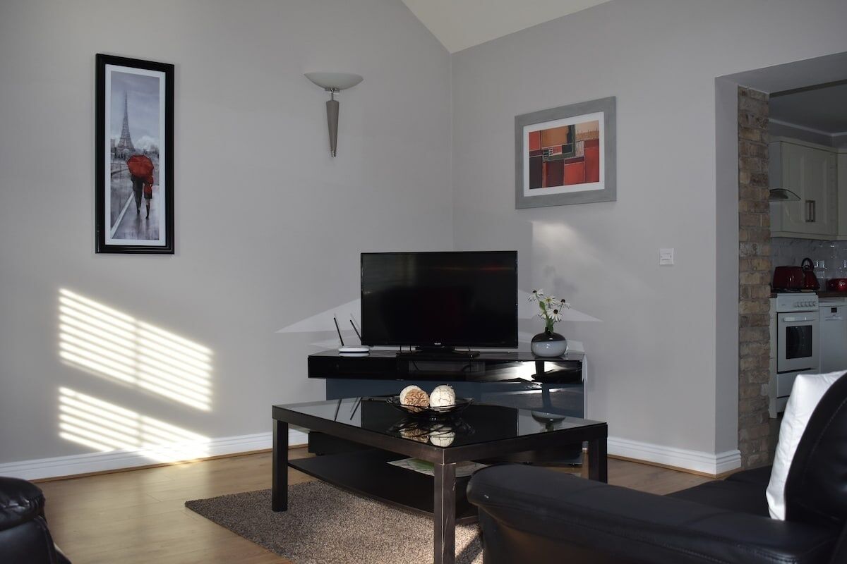 Ein modernes Wohnzimmer mit einem Flachbildfernseher, einem Couchtisch und einer Couch. An der Wand hängt ein gerahmtes Bild, und durch eine offene Tür ist eine Küche zu sehen.