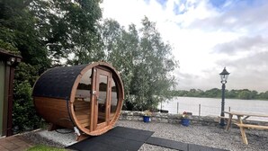 A wooden barrel-shaped structure with glass doors, a picnic table, and a lamp post by a lake.