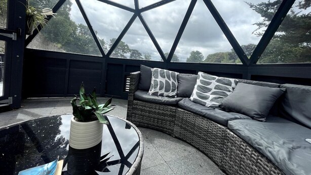 Un área de estar moderna al aire libre con techo de vidrio geométrico, un sofá de mimbre con cojines y una mesa de vidrio con una planta.