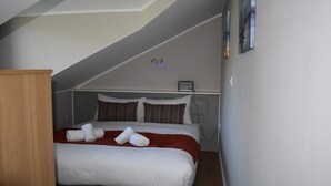 Un lit bien fait, avec une literie blanche et rouge, une armoire en bois et une petite étagère sur laquelle se trouve une lampe.