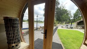 A wooden interior with a large circular window, a Harvia fireplace, and an outdoor view of a park with a picnic table and a stone structure.