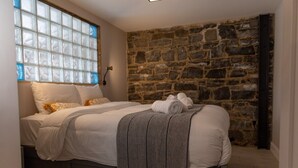 Un dormitorio con pared de piedra, un ventanal con persianas, cama con ropa blanca y una toalla enrollada sobre la cama.