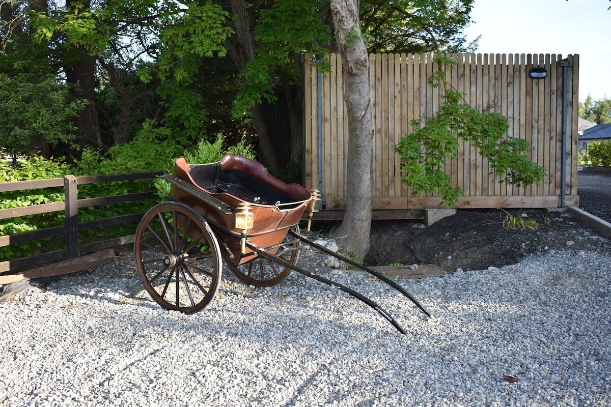 Une charrette en bois, dotée d’un siège en cuir et d’une longue barre, garée sur un chemin de gravier.