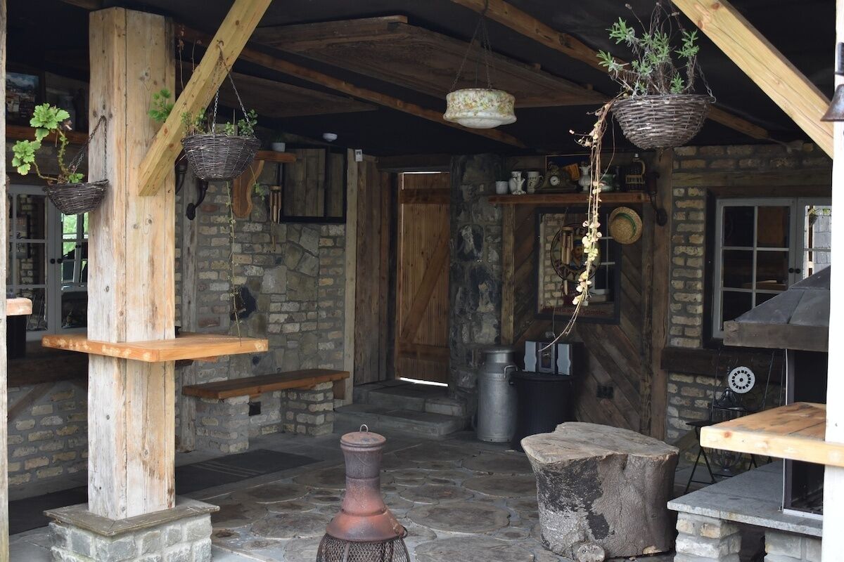 Un interior rústico con paredes de piedra, vigas de madera, cestas colgantes con plantas, una mesa de piedra y un horno de ladrillo.
