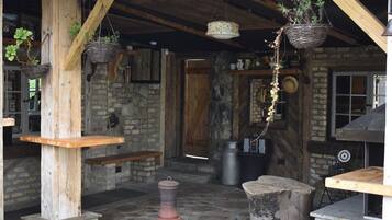 Un interior rústico con paredes de piedra, vigas de madera, cestas colgantes con plantas, una mesa de piedra y un horno de ladrillo.