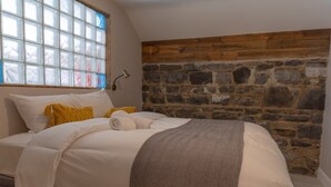 Une chambre avec un mur en pierre, un lit avec une literie blanche, un plaid jaune et une fenêtre à vitres dépolies.