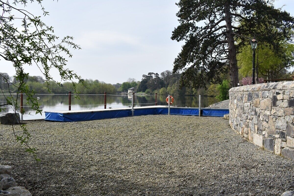 A pebbled waterfront area with a stone wall, a dock, and a calm lake in the background.