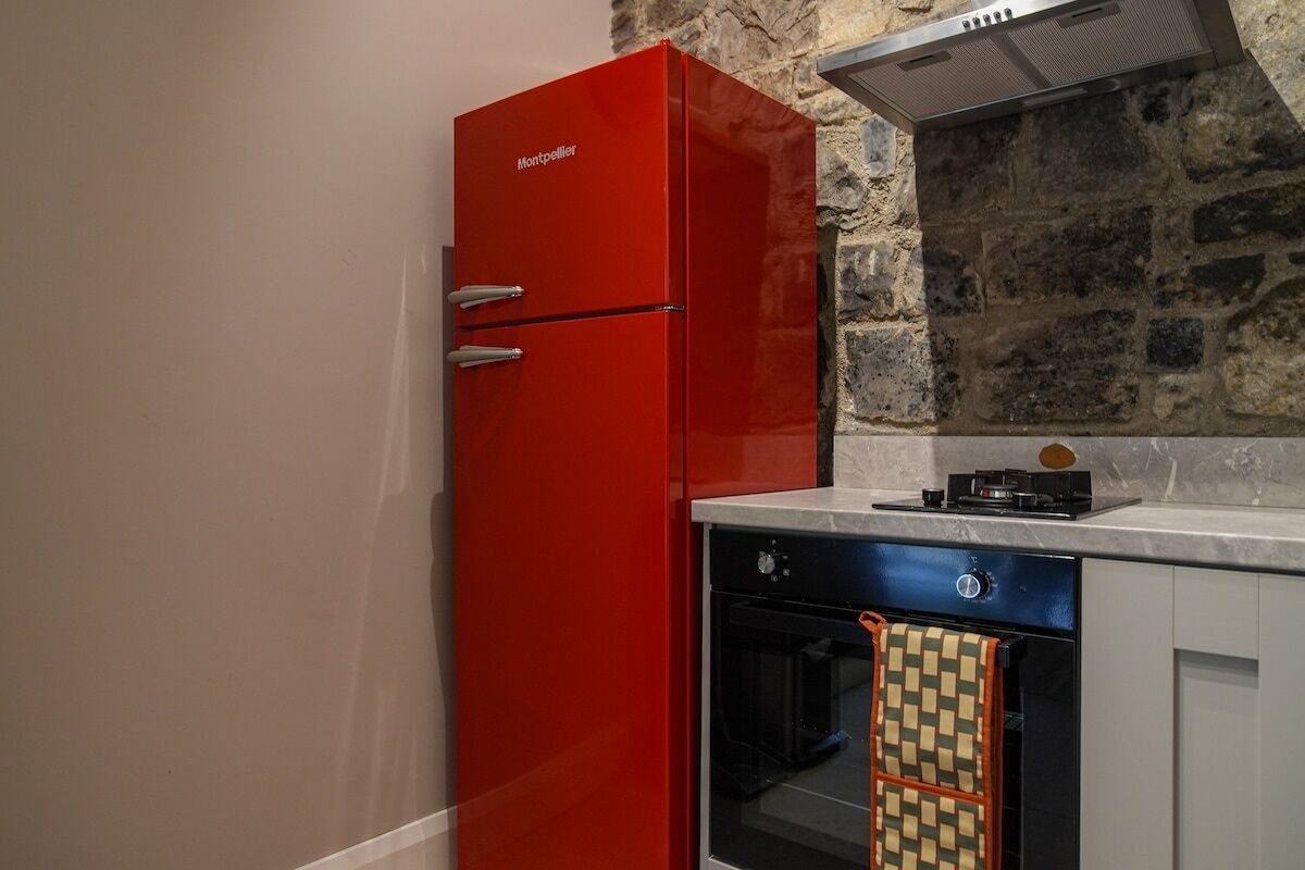 A compact kitchen with a red refrigerator, a built-in oven, and a stone wall backsplash.