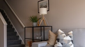 Una sala de estar con un sofá gris, cojines estampados, una mesita auxiliar negra con una planta, una lámpara de pie y un cuadro en la pared.