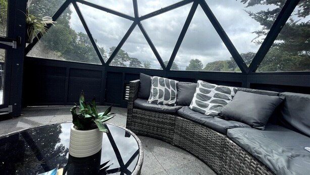 Un área de estar moderna al aire libre con techo de vidrio geométrico, un sofá de mimbre con cojines grises y una mesa de centro de vidrio con una planta.