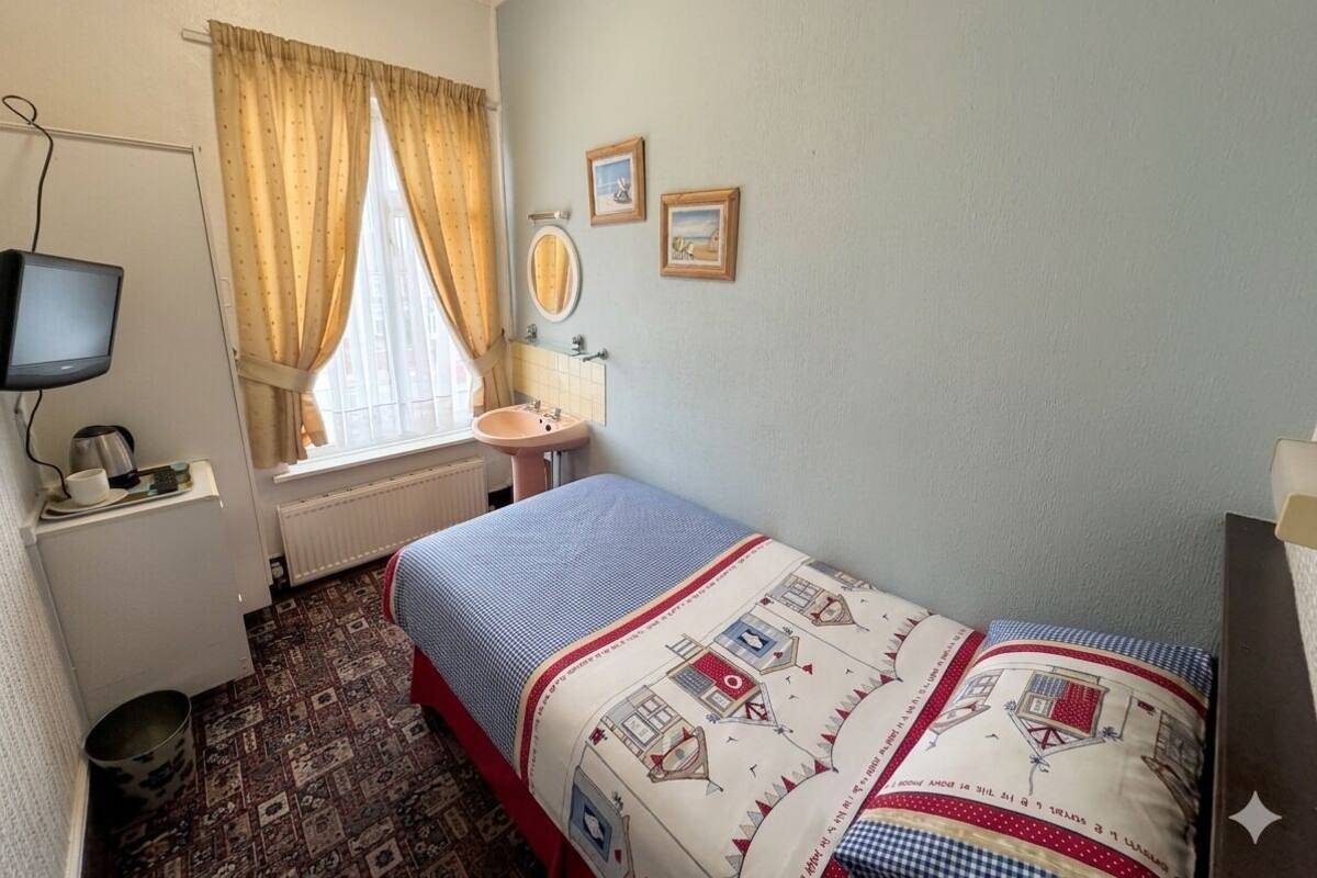 Um quarto pequeno com uma cama de solteiro, janela com cortinas, mesa de cabeceira com espelho, televisão e radiador.