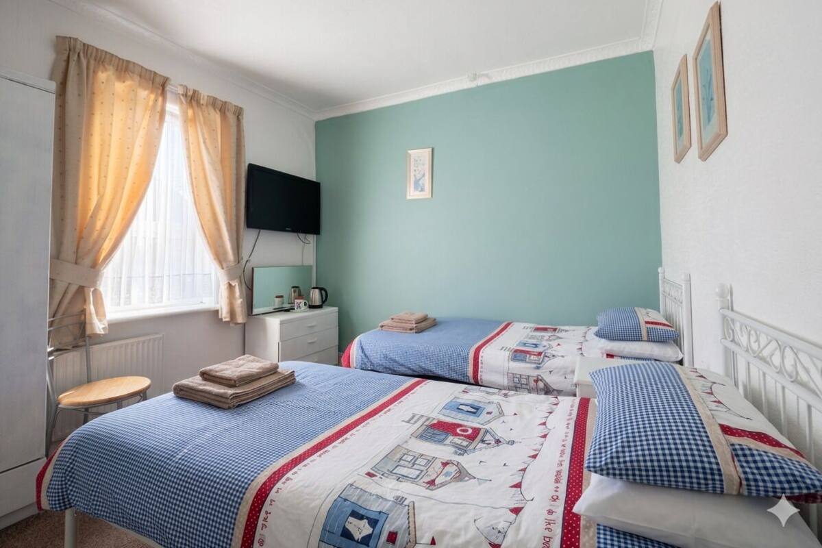 Quarto com duas camas, janela com cortinas, mesa de cabeceira e televisão fixada na parede.