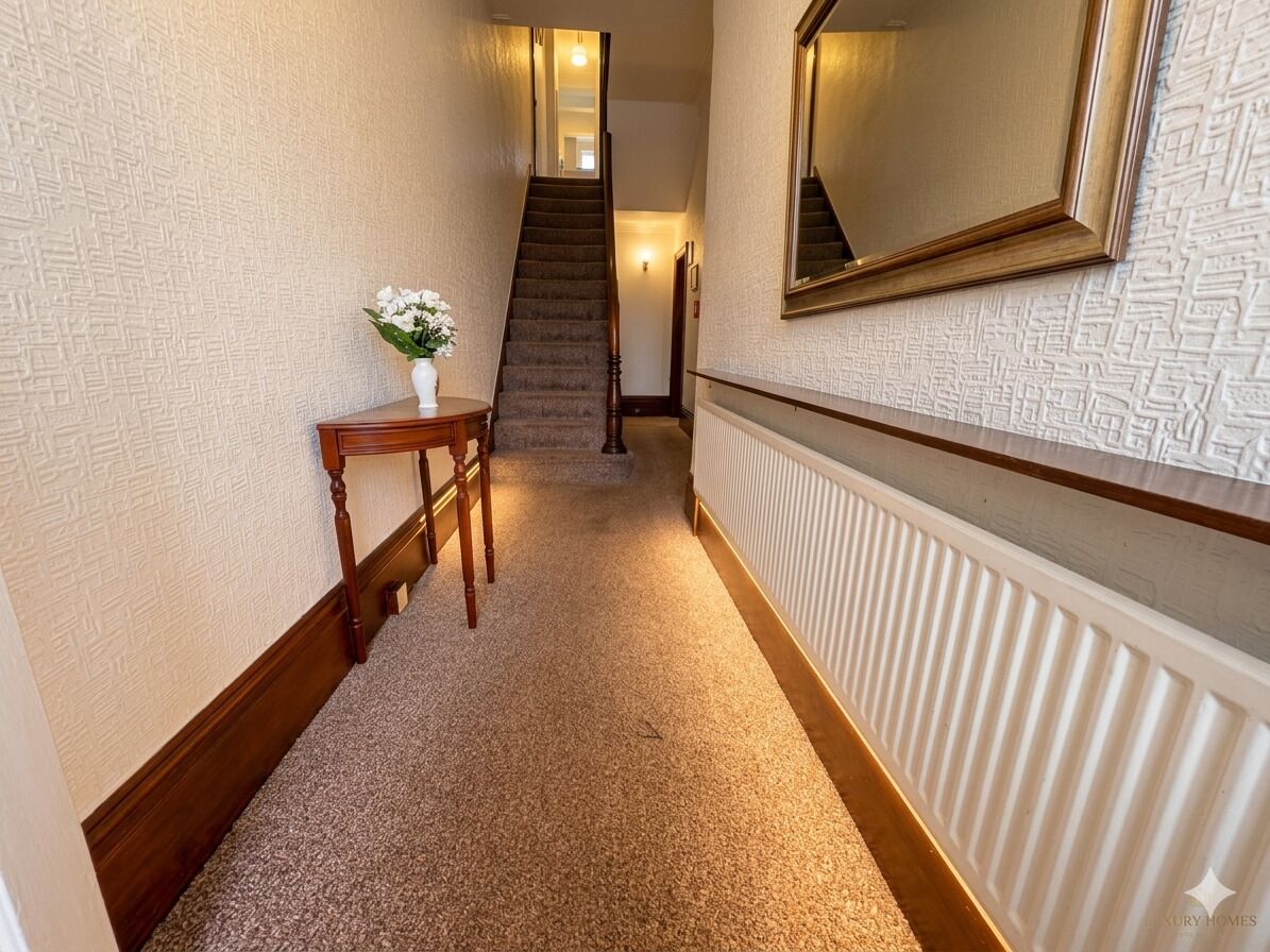 A carpeted hallway with a wooden table and vase, a staircase, and a wall-mounted mirror.