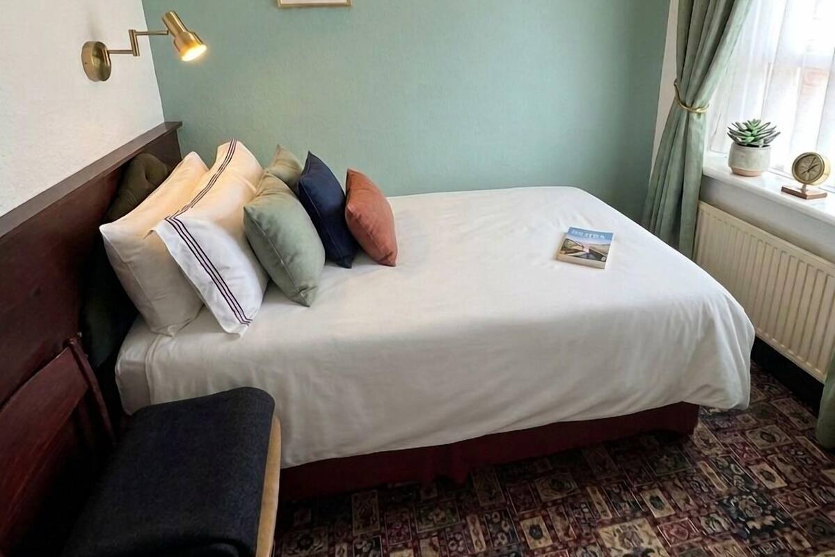 Quarto de hotel com uma cama, uma cadeira, uma luminária na parede e uma janela com cortinas.