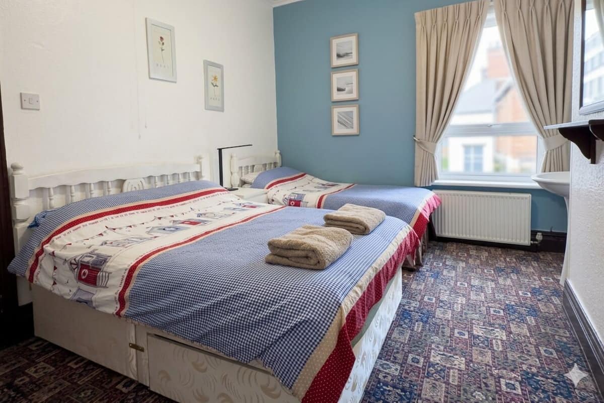 Uma cama de casal com colcha xadrez, duas mesas de cabeceira, um radiador e uma janela com cortinas.