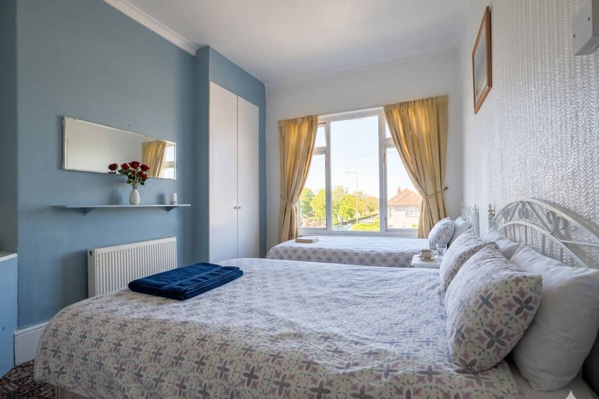 Quarto com duas camas, janela com cortinas, radiador, espelho e prateleira na parede com um vaso.