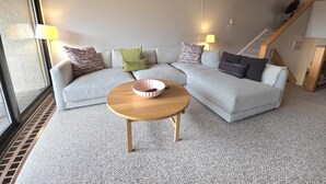 Ein modernes Wohnzimmer mit einem hellgrauen Ecksofa, einem runden Couchtisch aus Holz und einem großen Fenster, das natürliches Licht hereinlässt.