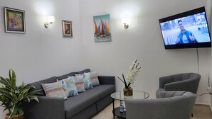 Ett modernt vardagsrum med en grå soffa, ett soffbord i glas och en grå fåtölj. Det finns två väggmonterade lampor, en platt-TV och en inramad tavla ovanför soffan.