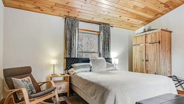 Ein Schlafzimmer mit Holzbalkendecke, einem Bett mit weißer und grauer Bettdecke, einem Schaukelstuhl, einem Nachttisch mit Lampe und einem Kleiderschrank.
