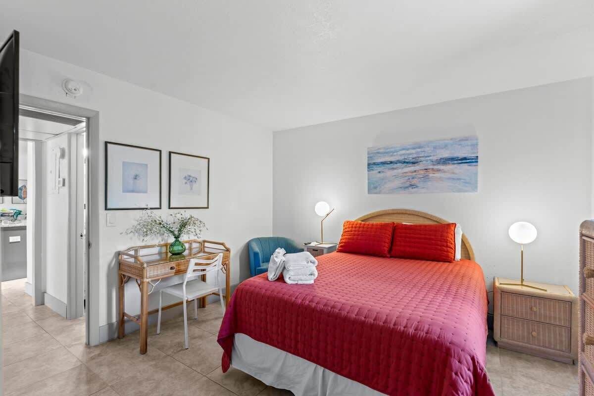 Habitación de hotel con cama, un escritorio con silla, una mesita con un florero y un cuadro en la pared.