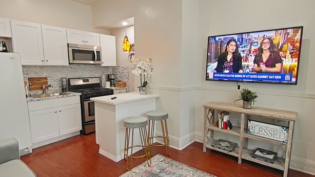 A modern kitchen with white cabinets, a central island, and a flat-screen TV displaying a news broadcast.