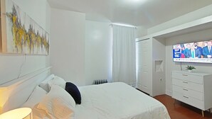 A bedroom with a bed, a TV, a radiator, a white wardrobe, and a painting on the wall.