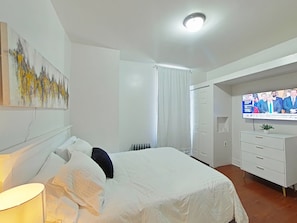 A bedroom with a bed, a TV, a radiator, a white wardrobe, and a painting on the wall.