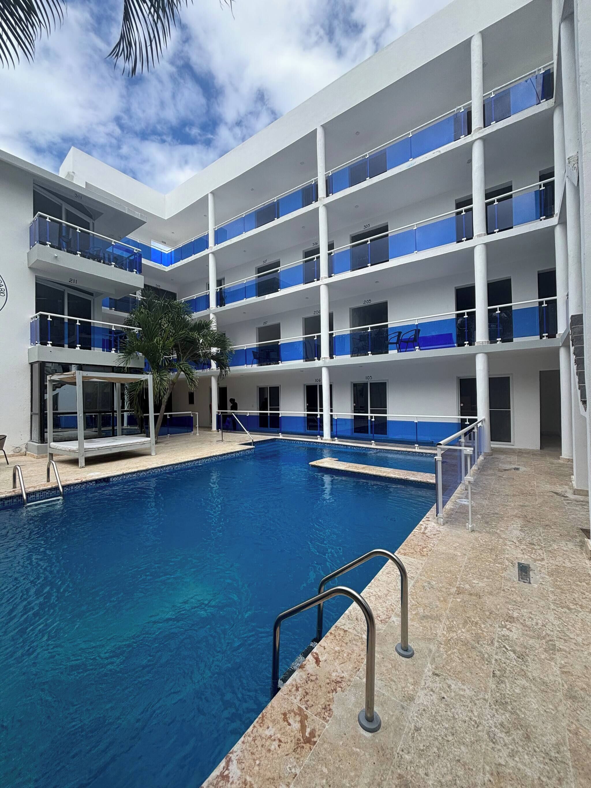 A multi-story hotel with a central swimming pool, balconies with blue railings, and a palm tree.