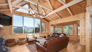 A spacious cabin with wooden walls and ceilings, large windows offering scenic mountain views, a leather sofa, and a stone fireplace. - Bass Lake luxury cabin with lake views & boat slip (Bass Lake)