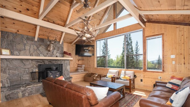 A cozy cabin interior with a stone fireplace, leather sofas, and large windows offering a view of the forest. - Bass Lake luxury cabin with lake views & boat slip (Bass Lake)