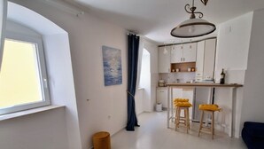 A modern kitchen with white cabinets, a bar area, and stools. There is a window on the left and a painting on the wall.