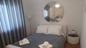 Un lit bien fait, recouvert d’une couvre-lit à motifs bleus et agrémenté d’oreillers blancs, un miroir rond accroché au mur, une petite table de chevet avec une lampe et une poubelle.