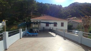 Une terrasse extérieure couverte, dotée d’une table et de chaises, entourée d’une clôture blanche et offrant une vue sur une colline boisée.