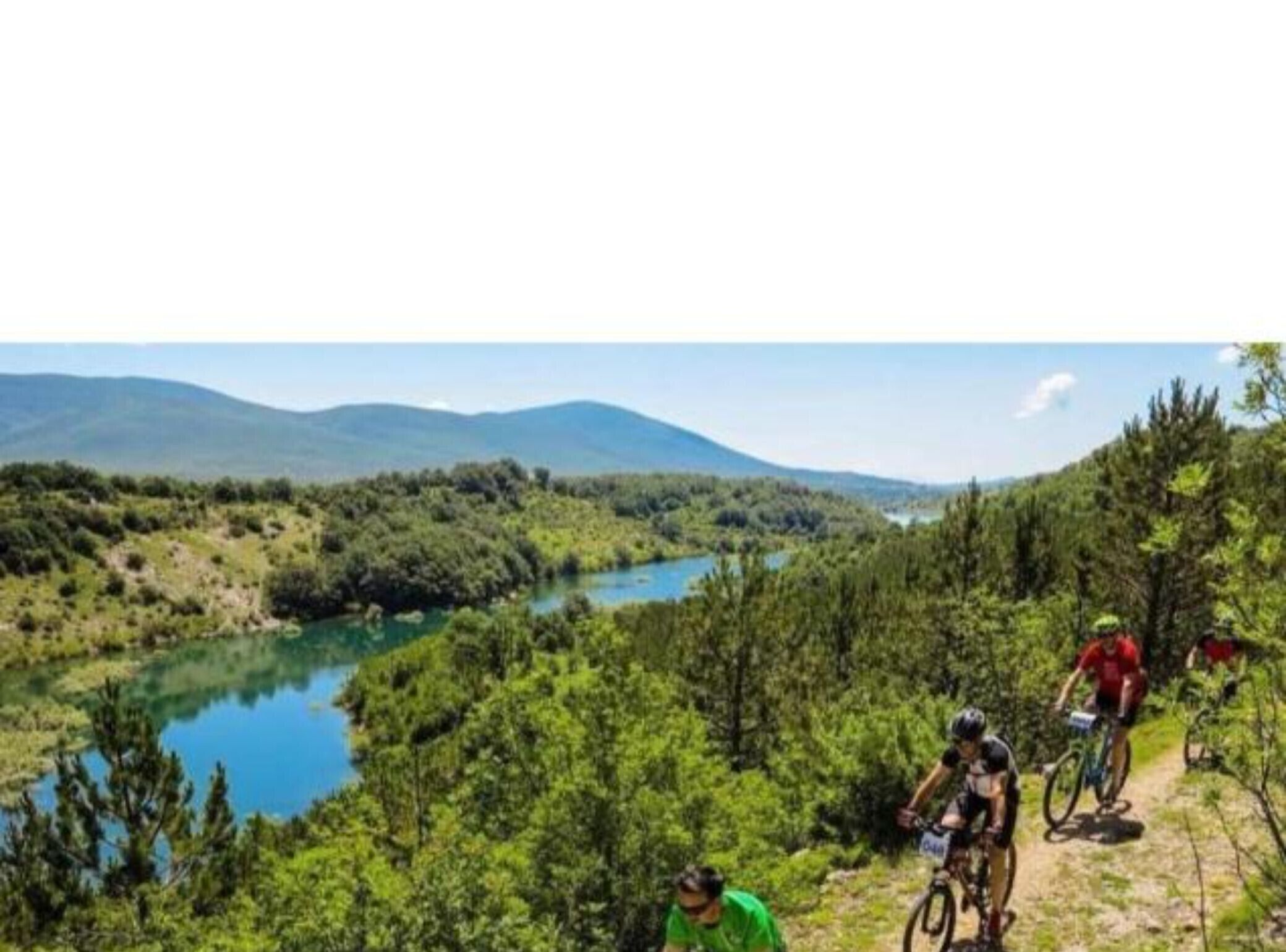 Un paysage montagneux avec une rivière, des cyclistes sur un sentier et une forêt dense.